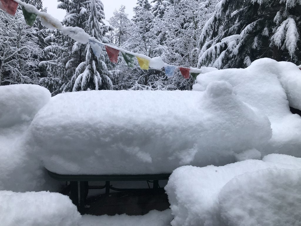 Outdoor furniture buried under mountains of snow, prayer flags and snow-covered trees in the background